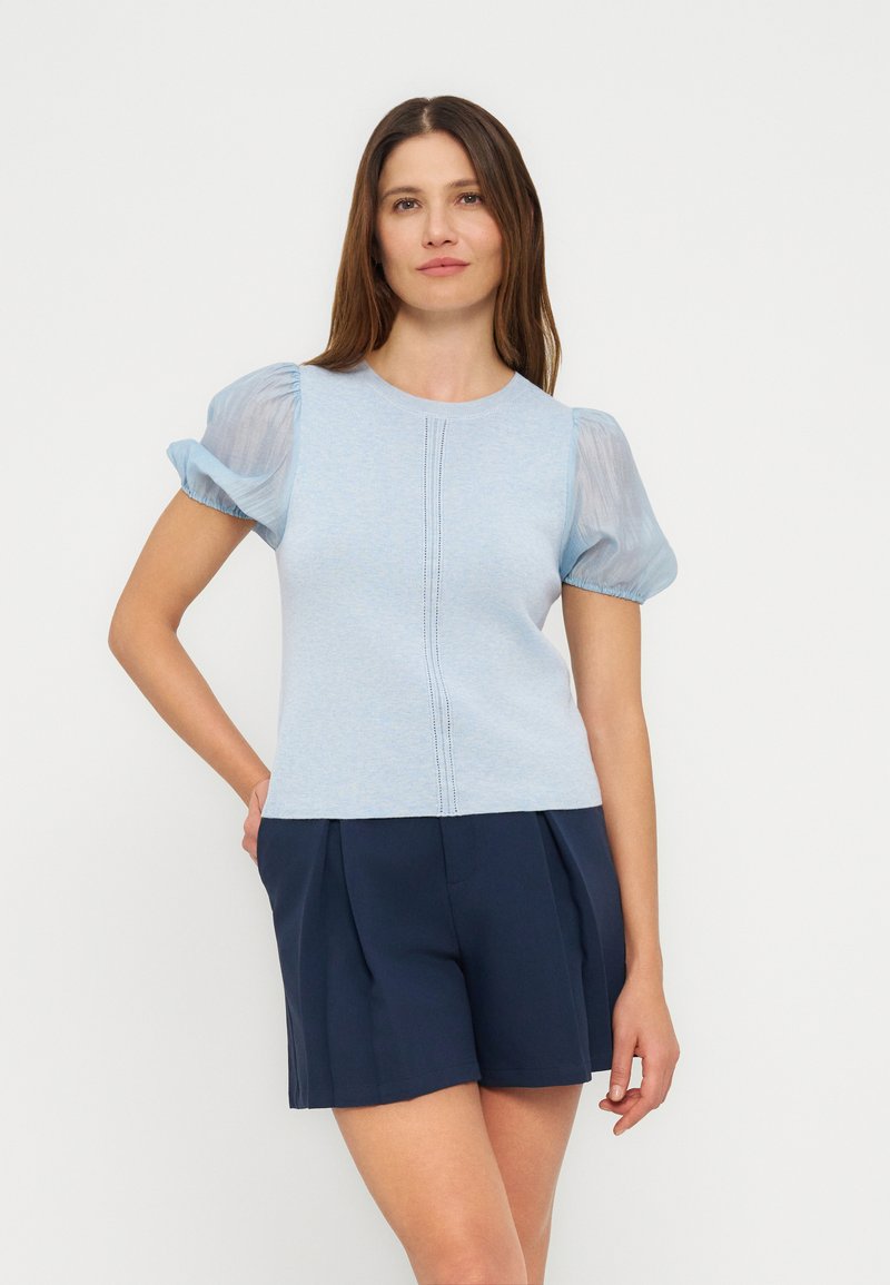 Γυναίκα που στέκεται φορώντας ανοιχτό γαλάζιο τοπ με διαφανείς φουσκωτές μανίκες και σκούρα μπλε ψηλόμεσα σορτς με πιέτες, μπροστά σε απλό λευκό φόντο.