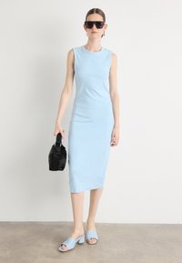 Světle modré midi šaty bodycon bez rukávů, vyrobené z jemné látky, s malým logem. Kombinováno se světle modrými nazouvacími botami a černou kabelkou.