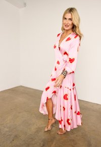 Ξανθιά γυναίκα με ροζ φόρεμα τύπου wrap με κόκκινα μοτίβα καρδιάς και ψηλοτάκουνα σανδάλια με λεοπάρ σχέδιο, στέκεται σε τσιμεντένιο πάτωμα μπροστά από λευκό τοίχο.