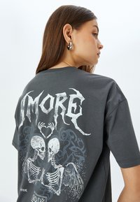 Camiseta de algodón gris con un gráfico de dos esqueletos con alas y un corazón, texto "AMORE" en una fuente blanca desgastada en la parte posterior.