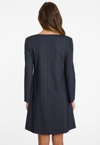 Frau mit langen braunen Haaren trägt ein knielanges, marineblaues, gestreiftes Kleid mit langen Ärmeln, von hinten auf einem weißen Hintergrund gezeigt.