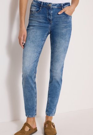 Kvinde iført lyseblå slim-fit jeans og brune ruskind slip-on sko, stående mod en ensfarvet lys baggrund.