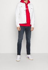 Witte denimjas over een rode hoodie, donkere jeans en witte sneakers met blauwe en rode accenten. Eenvoudig ontwerp met een casual pasvorm.