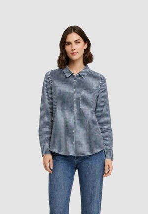 Donna che indossa una camicia a righe verticali blu e bianche con bottoni e jeans blu, in piedi davanti a uno sfondo grigio chiaro uniforme.