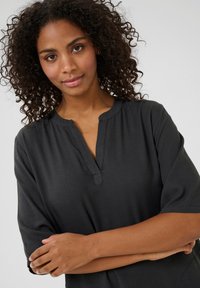 Haut noir à manches courtes avec un col rond et un petit décolleté en V. Texture lisse avec une coupe décontractée, porté par un mannequin avec des cheveux bouclés.