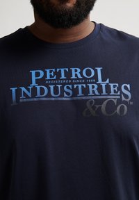 Náměsíčně modré bavlněné tričko s předním grafickým textem: "PETROL INDUSTRIES" v různých odstínech modré a šedé, s "& Co" na spodní části.
