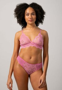 Vaaleanpunainen pitsinen bralette, jossa on säädettävät hihnat ja aaltoilevat reunat, yhdistettynä yhteensopiviin bikinialushousuihin, joissa on läpikuultavia paneeleita ja pitsiä.
