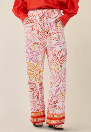 Weiße Hosen mit weitem Bein, bedruckt mit tropischen Blättern in Rosa, Rot und Orange, Kordelzug in der Taille und gemustertem Saum, getragen mit roter Bluse und Sandalen.