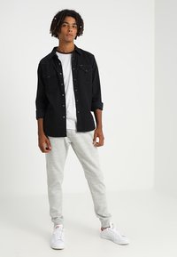 Camisa negra de botonadura con dos bolsillos en el pecho, camiseta blanca de cuello redondo, pantalones de chándal gris claro y zapatillas blancas con detalles rojos.