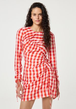 Vrouw met lang krullend haar die een rood-wit geruite, geplooide minijurk met lange mouwen en trekkoorden draagt.