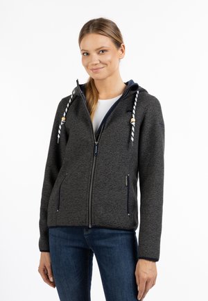 Giovane donna che indossa una felpa con cappuccio grigio scuro con zip e cordini a righe, e jeans blu, sorridendo leggermente su sfondo bianco.