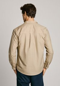 Camicia beige con bottoni, dotata di una piega sulla schiena, maniche lunghe e polsini con bottoni. Presenta un colletto classico e un tessuto liscio e testurizzato.