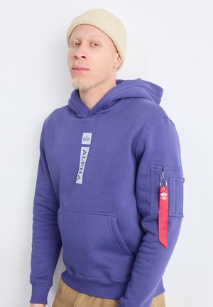 Lila Kapuzenpullover mit einer vorderen Kängurutasche, ausgestattet mit einem Logo auf der Brust und einer mit Reißverschluss versehenen Armtasche mit einem roten Ziehband.