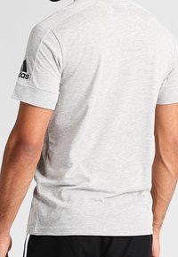 Maglietta sportiva grigia realizzata in tessuto misto cotone, con maniche corte e logo Adidas sulla manica sinistra. Texture liscia.