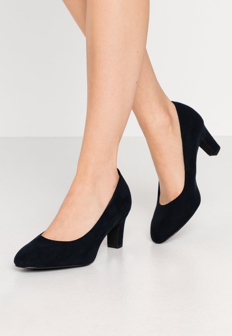 Femme portant des escarpins en daim noir à talon moyen, debout avec un pied légèrement levé sur un sol gris clair et un fond blanc.