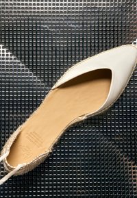 Chaussure plate espadrille beige avec bout pointu fermé et talon tressé, reposant sur une surface texturée noire présentant des perforations carrées.