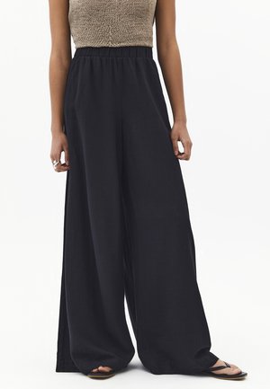 Femme portant un pantalon noir taille haute à jambes larges, un haut en maille beige sans manches, des bagues à la main gauche et des tongs noires sur fond blanc.