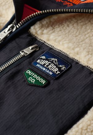 Marineblaue Stoff mit strukturierter Oberfläche, gefüttert mit beigem Fleece und drei gestickten Aufnähern: Berge, Logo eines Outdoor-Unternehmens.