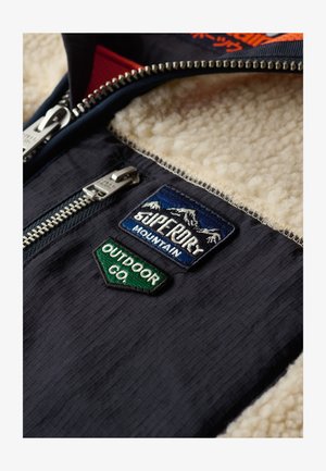 Granatowy materiał o teksturowanym wykończeniu, z beżowym futrem od wewnątrz oraz trzema haftowanymi naszywkami: góry, logo firmy outdoorowej.