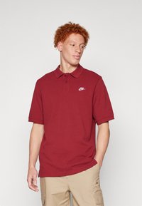 Μπορντό πόλο μπλουζάκι με κοντά μανίκια, διαθέτει πατιλέτα με τρία κουμπιά και λευκό λογότυπο Nike στο αριστερό στήθος. Υλικό από μείγμα βαμβακιού.