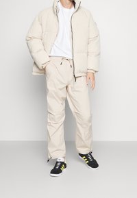 Beige gesteppte Jacke mit hohem Kragen, kombiniert mit hellbeigen Hosen und schwarzen Turnschuhen mit gelben Streifen. Schlichtes Design mit minimalen Akzenten.