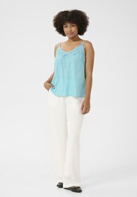 Šviesiai mėlyna camisole su nėriniu apykaklėje, derinama su baltais plačiakelniais kelnais ir juodais avalynės. Vienoda balta fono spalva.