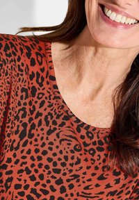 Leopard print top in rust color, featuring a wide neckline, soft fabric, and black spots in varying sizes. Smooth texture and casual fit.