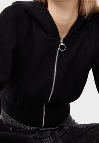 Felpa nera con zip in morbido tessuto, dotata di cappuccio, orlo a coste e dettaglio cerniera argento, con una vestibilità leggermente sagomata.