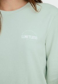 Lys grønn sweatshirt med rund hals. Har et hvitt trykt logo "LIMITLESS" og teksten "ONLY PLAY ENERGY" over logoen.