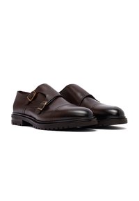 Een paar donkerbruine leren double monk strap herenschoenen met ronde neuzen en dikke rubberen zolen op een witte achtergrond.
