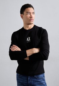 Black long-sleeve t-shirt with a white logo on the chest, paired with blue denim jeans. The fabric appears smooth with a fitted design.