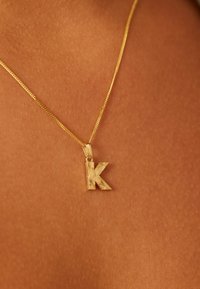 Gouden hangerketting met een gestructureerde letter "K" bedel aan een dunne ketting, gelegen op een huidkleurige achtergrond.