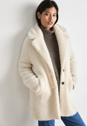Cream-colored, fuzzy coat with large lapels, two buttons, and side pockets. Worn over a brown turtleneck and blue jeans.