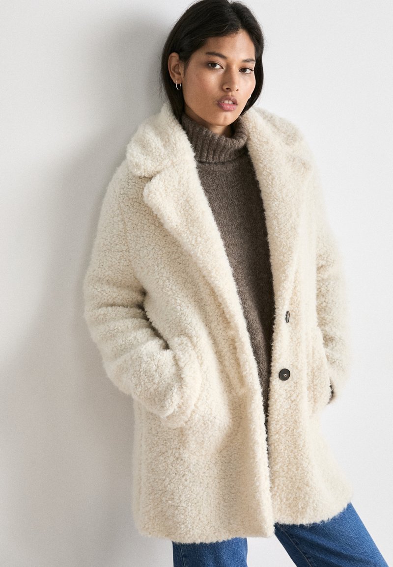 Cream-colored, fuzzy coat with large lapels, two buttons, and side pockets. Worn over a brown turtleneck and blue jeans.
