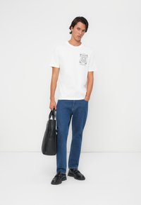 T-shirt en coton blanc avec un design de poche graphique, associé à un jean en denim bleu. Le modèle tient un sac fourre-tout en cuir noir, portant des chaussures noires à enfiler.