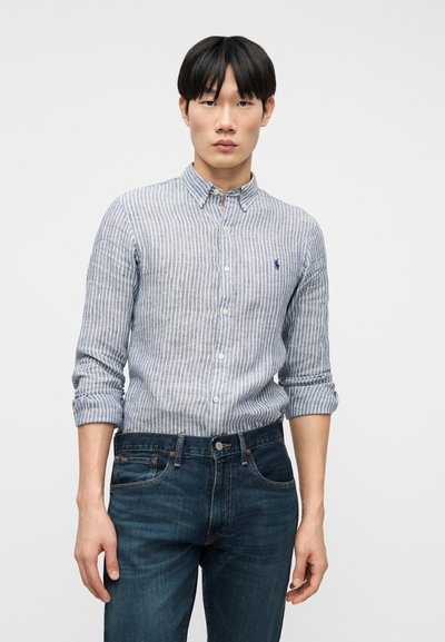 Νέος άνδρας που φοράει μπλε και λευκό πουκάμισο με κάθετες ρίγες και μανίκια γυρισμένα, καθώς και σκούρο μπλε τζιν, μπροστά σε απλό φόντο.