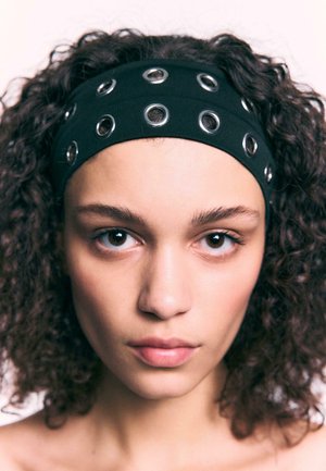 Bandeau en tissu noir avec accents en œillets argentés, surface texturée et design élastique, porté par un modèle aux cheveux bouclés.