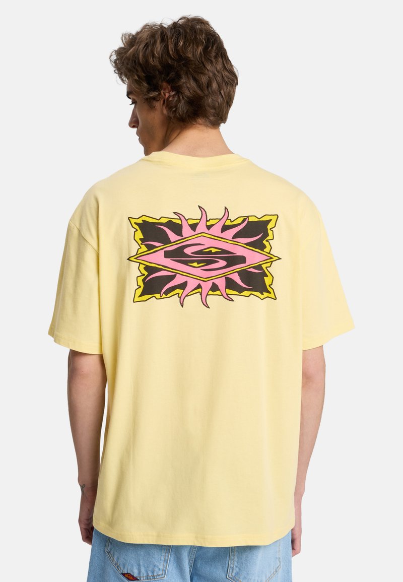 Homme aux cheveux bouclés portant un T-shirt jaune surdimensionné avec un motif de soleil abstrait rose et noir dans le dos, associé à un jean bleu clair.