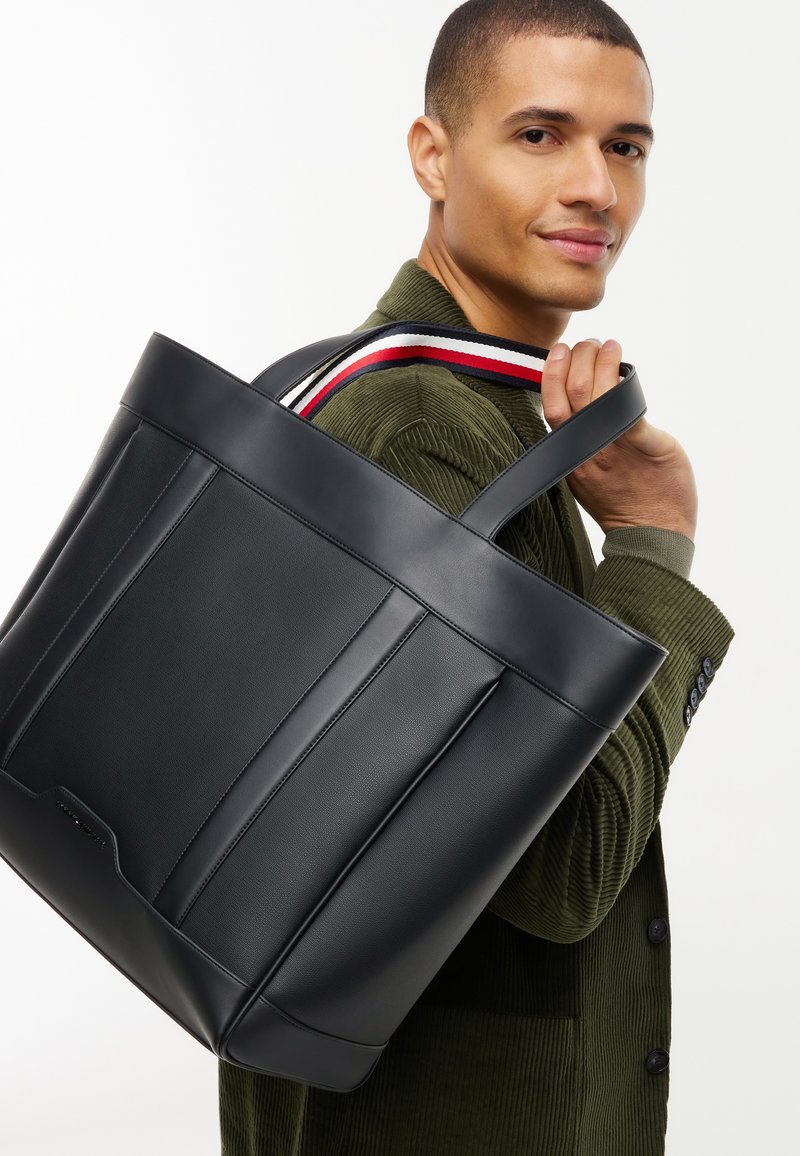 Homme en veste en velours côtelé verte portant un grand sac cabas en cuir noir avec une bandoulière rayée rouge, blanche et bleue.