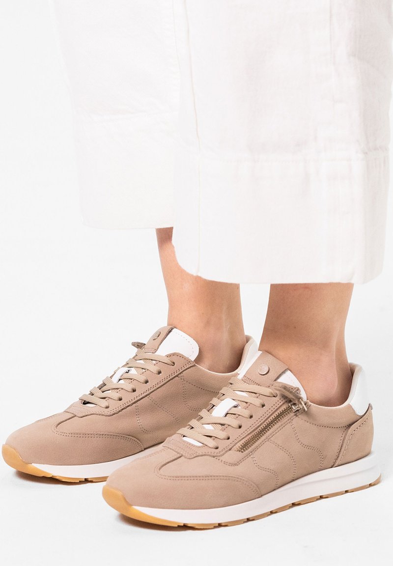 Beige sneaker van suede met witte accenten, ronde neus, vetersluiting en zijritsdetail. Rubberen zool met een gestructureerde grip.