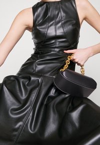 Svart läderklänning med figurnära bodice och draperad kjol. Håller en liten oval svart väska med guldkedjehandtag och vita söm detaljer.