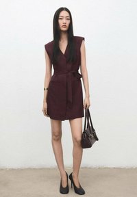 Bordowa sukienka bez rękawów z dekoltem w serek, z paskiem w talii i bocznymi kieszeniami, zestawiona z czarnymi szpilkami i małą ciemną torebką. Gładka tkanina.