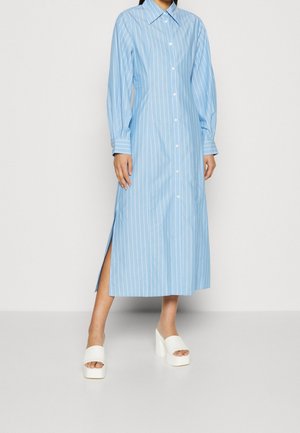 Kvinde iført en lang blåstribet kjole med knapper og lange ærmer samt hvide platformsandaler, stående på en grå gulv.