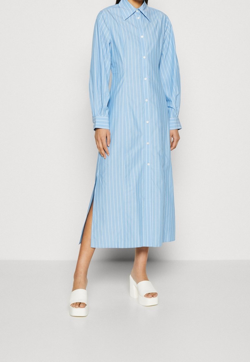 Femme portant une longue robe chemise à rayures bleues avec manches longues et sandales à plateforme blanches, debout sur un sol gris.