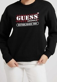Sudadera negra con un diseño de cuadrícula roja y un logo en blanco que dice "GUESS ORIGINALS ESTABLISHED 1981". Puños y dobladillo acanalados.