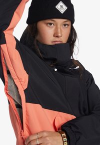 Bicolorová bunda s čiernym telom a koralovými akcentmi, vyrobená z ľahkého materiálu. Obsahuje vysoký golier a zipsové vrecká.