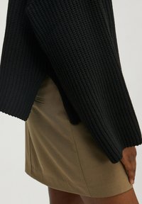 Schwarzer Strickpullover mit einem strukturierten Muster und geripptenÄrmeln. Kombiniert mit einem khakifarbenen Minirock aus glattem Stoff und dezenten Seitennähten.