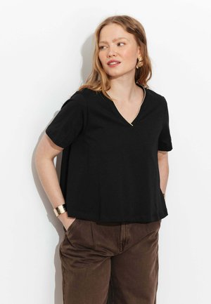 Femme aux cheveux longs portant un haut noir à manches courtes, un pantalon marron, des boucles d'oreilles en or et un large bracelet en or, regardant vers sa gauche.