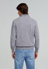 Grauer Pullover mit hohem Kragen, gerippten Bündchen und Saum; weiche Textur. Kombiniert mit hellblauen Jeans.