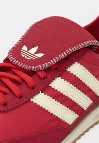 Scarpa da corsa rossa in materiali di suede e tessuto. Presenta un design con tre strisce bianche, cuciture decorative e un'etichetta con il marchio sulla linguetta.